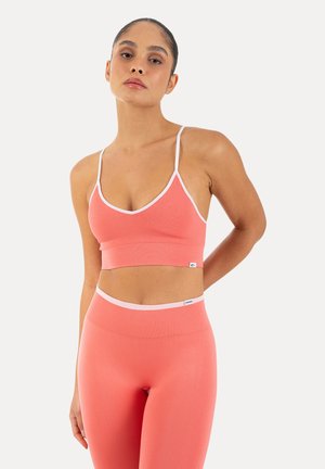 Mujer con el pelo recogido que lleva un sujetador deportivo y mallas sin costuras color coral con ribete blanco, de pie frente a un fondo claro y liso.