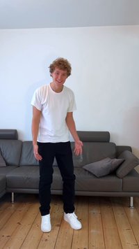 Grey leather sofa with two patterned cushions, wooden floor, person in a white t-shirt, black pants, and white sneakers, standing indoors.