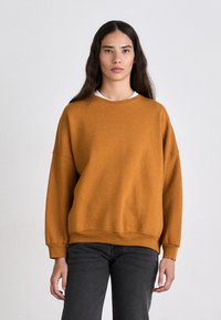 Sudadera de gran tamaño color mostaza con cuello redondo, puños y dobladillo acanalados. Textura suave, mostrada con jeans de color gris oscuro.