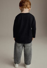Chemise noire à manches longues avec col rond, associée à un jean ample gris clair. Baskets blanches visibles aux pieds.