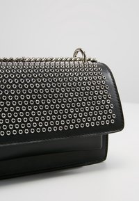 Sac à main en cuir noir avec des œillets métalliques argentés recouvrant le rabat et une bandoulière en chaîne argentée sur un fond clair uni.