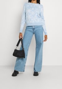 Hellblaues Sweatshirt mit weißem Schneeflockenmuster, kombiniert mit hellblauen, weiten Jeans. Hält eine schwarze Handtasche aus Stoff mit verstellbarem Tragegurt.