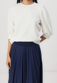 Sudadera blanca, texturizada, de cuello redondo con mangas abullonadas, combinada con una falda de tablas azul marino con una cintura ancha.