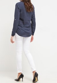 Kvinna klädd i en navyblå mönstrad långärmad skjorta, vita skinny jeans och svarta högklackade skor, vänd bort från kameran mot en vit bakgrund.