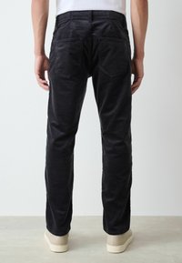 Black corduroy trousers with a straight cut, featuring two back pockets and a smooth texture. Paired with beige sneakers.