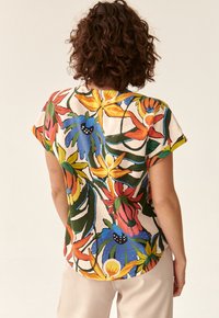 Kortærmet blomstret bluse med livlige farver (rød, blå, gul) på en lys baggrund; har en afslappet pasform og buet kant.