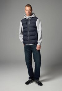 Gilet rembourré bleu marine avec une capuche grise, des manches grises et une fermeture éclair à l'avant. Un jean foncé et des chaussures de sport noires complètent la tenue.