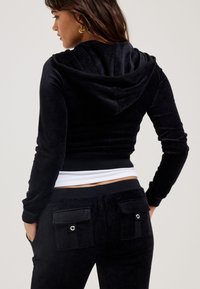 Black velvet zip-up hoodie with a hood, paired with black pants featuring front pockets and snap-button details. Smooth texture throughout.
