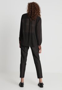 Schwarze, halbtransparente Bluse mit weißen Punkten, langen Ärmeln und einem bedruckten, gemusterten Kragen, kombiniert mit schwarzen Nadelstreifen-Hosen.