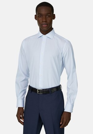 Mężczyzna ubrany w jasnoniebieską koszulę w paski z długim rękawem, wpuszczoną w ciemnoniebieskie spodnie z czarnym paskiem, stojący na jednolitym tle.