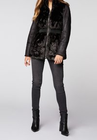 Czarna kurtka z futra syntetycznego z skórzanymi rękawami, zamek błyskawiczny i ściągacz w talii. W zestawie z ciemnymi, obcisłymi dżinsami i czarnymi botkami na obcasie.