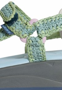 Sandalias tejidas con tiras azules y verdes de textura trenzada, aseguradas por un anillo de plástico rosa, sobre una suela de color azul claro.
