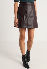 Burgunderrote Leder-Minirock mit einer glatten Textur, mit einem taillierten Bund und dezenten Steppnähten. Kombiniert mit schwarzen Stiefeletten.