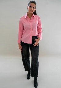 Roze overhemd met knopen en lange mouwen, zwarte high-waisted jeans en zwarte enkellaarzen. Het overhemd heeft een gladde textuur en een aansluitende snit.