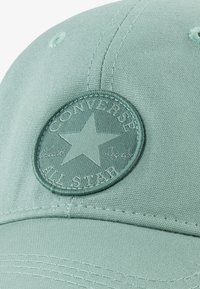 Mētas zaļā kokvilnas cepure ar apaļu reljefu plāksteri, uz kura redzams zvaigzne un uzraksts "CONVERSE ALL STAR"; teksturēts apdare un izšūtas detaļas.