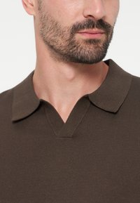 Homme barbu portant un polo en maille marron avec un col et une encolure en V, montré du cou à la partie supérieure de la poitrine.