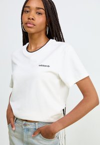Hvit bomulls t-skjorte med korte ermer, svart logo på brystet, og svart kant rundt halsåpningen. Kombinert med lyseblå denimshorts.