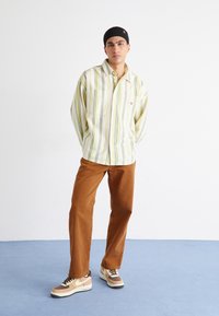 Camisa de rayas de manga larga y con botones en amarillo claro, verde y blanco; combinada con pantalones marrones y zapatillas marrones y beige con un swoosh blanco.
