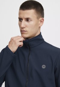 Mornarsko modra zip-up jakna iz mehkega materiala, z visokim ovratnikom in okroglo logotipom na levi strani prsnega dela. Gladka tekstura z oprijetim krojem.