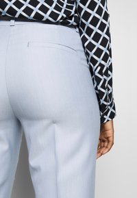 Un pantalon bleu clair en tissu lisse, doté d'une poche arrière. Un haut noir et blanc à motifs avec des designs géométriques est partiellement visible.