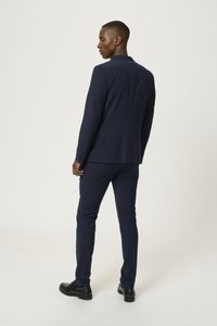 Costume bleu marine avec une coupe ajustée, une texture lisse et des revers en pointe. Des chaussures habillées noires complètent le look formel.
