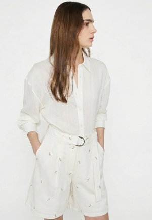 Joven con el pelo largo y castaño, que lleva una camisa blanca abotonada y pantalones cortos blancos de cintura alta con pequeños diseños bordados y un cinturón.