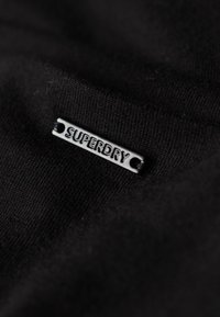 Czarna tkanina o teksturowym wykończeniu z srebrną metką z wytłoczonym napisem "SUPERDRY". Metka przymocowana za pomocą dwóch otworów.