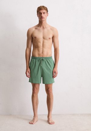 Junger Mann, barfuß gegen eine weiße Wand stehend, tragend eine grüne Shorts mit Kordelzug, Arme entspannt an den Seiten, neutrale Ausdruck, nach vorne gerichtet.
