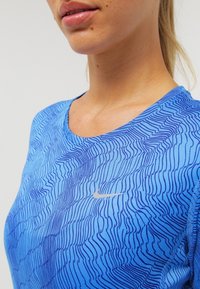 Sininen urheilupaita, jossa on aaltoileva viivakuviota, lyhyet hihat, pyöreä kaula-aukko ja pieni hopeinen Nike-logo edessä. Pehmeää ja venyvää materiaalia.