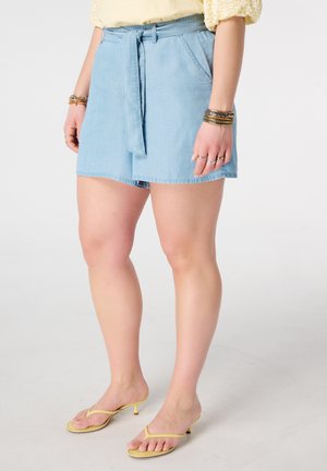 Femme portant un short en jean ceinturé bleu clair, des sandales à talons jaunes, un haut jaune et des bracelets aux deux poignets.