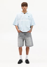 Polo shirt oversize azzurro chiaro con design stampato e testo, abbinato a pantaloni corti grigi con motivo e scarpe nere con lacci.