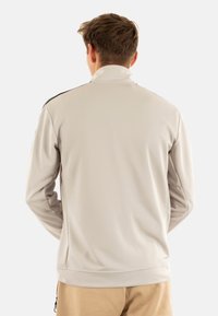 Chaqueta gris claro con un cuello alto, mangas largas y una textura suave. Presenta acentos negros en los hombros y un diseño ajustado.