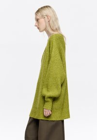 Oversized sweter z dzianiny, limonkowy, teksturowany materiał, opadające ramiona, długie rękawy, boczne rozcięcia, noszony na ciemnych spodniach.