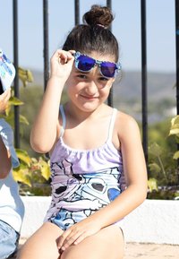 Maillot de bain aux couleurs violettes, bleues et blanches avec un design graphique. Enfant portant des lunettes de soleil bleues avec des accents floraux, assis en extérieur.