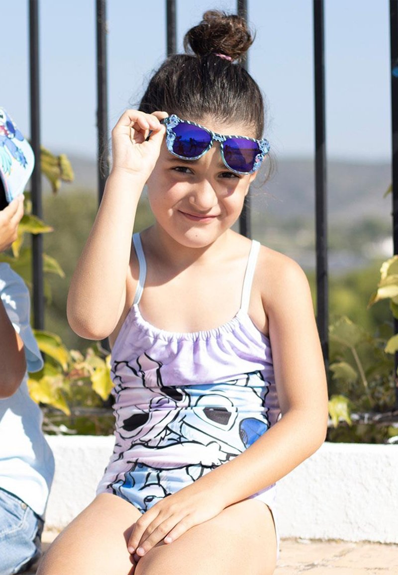 Maillot de bain aux couleurs violettes, bleues et blanches avec un design graphique. Enfant portant des lunettes de soleil bleues avec des accents floraux, assis en extérieur.