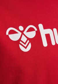 Tessuto di cotone rosso che mostra un grande logo bianco di un'ape stilizzata con elementi circolari e angolari, accompagnato dalla parola "hu."