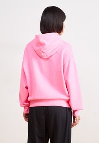 Felpa rosa con cappuccio e vestibilità rilassata, polsini e vita a coste. Tessuto in cotone liscio, vista da dietro, abbinata a pantaloni neri.