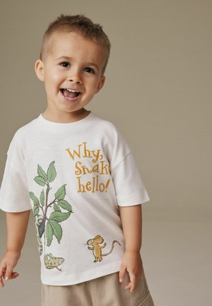 Jeune garçon souriant portant un T-shirt blanc avec un serpent, une souris et le texte « Pourquoi, bonjour serpent ! » sur un fond uni.