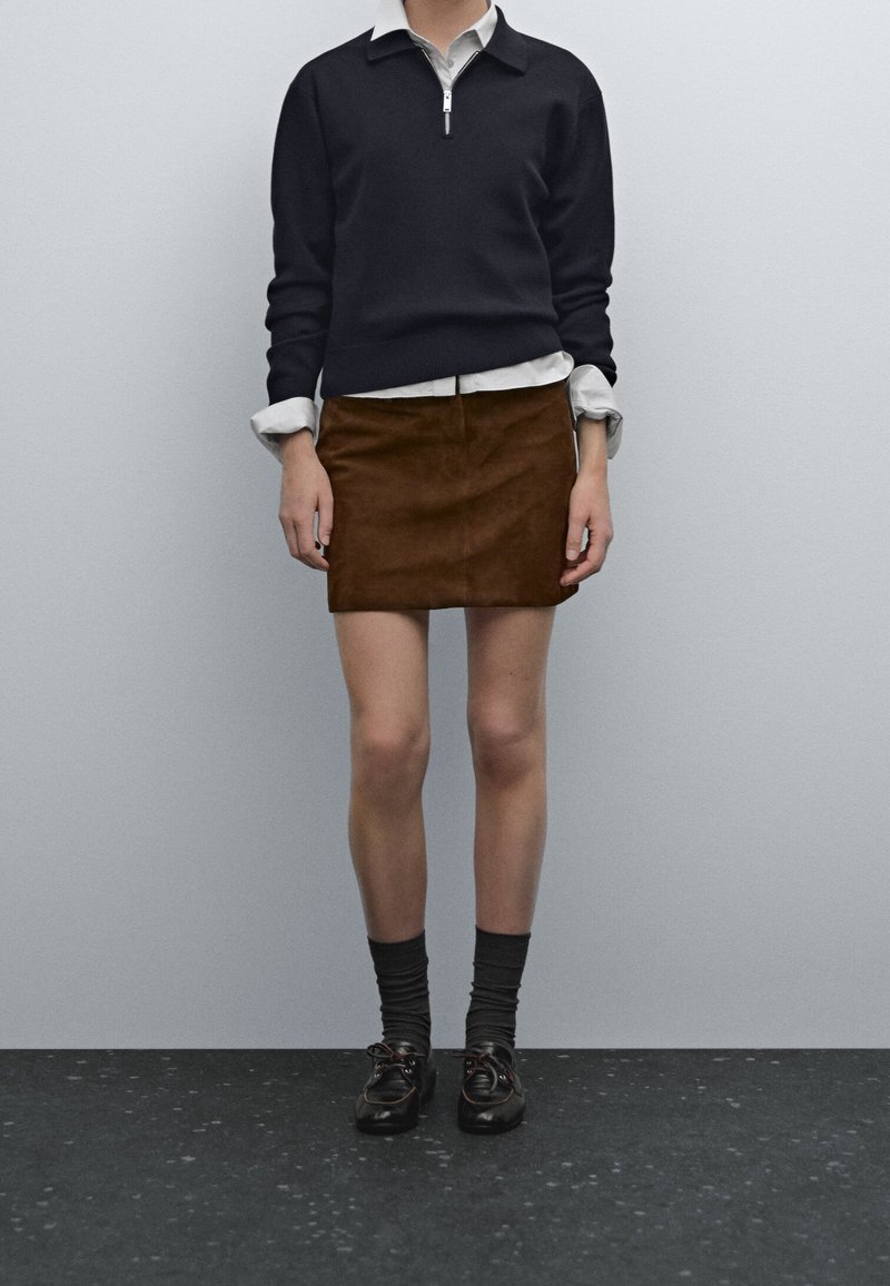 Marinblå zip-up-tröja, vit skjorta under, brun mocka minikjol, mörkgrå strumpor och svarta snörskor.