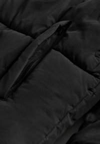 Nærbillede af en sort polstret jakke med synlig lynlås, kviltet tekstur og blød mat finish, der fremhæver materialet og syningens detaljer.