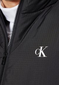 Schwarze gesteppte Reißverschlussjacke mit weißem "cK"-Logo auf der linken Brust. Der Stoff wirkt glatt mit einem strukturierten Karomuster.
