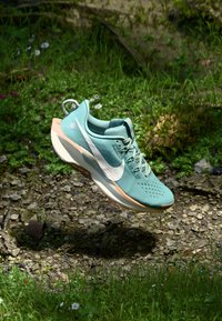 Light teal running shoe with a textured mesh upper, white swoosh logo, and peach-colored sole, floating above green grass and stones.