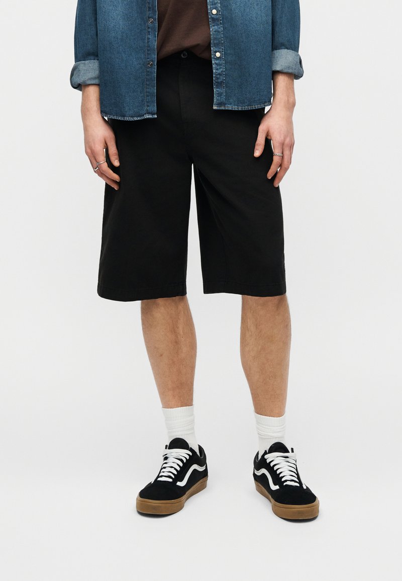 Personne portant un short noir jusqu'aux genoux, des chaussettes blanches, des baskets noires et blanches, une chemise en jean avec les manches retroussées, debout devant un fond blanc.