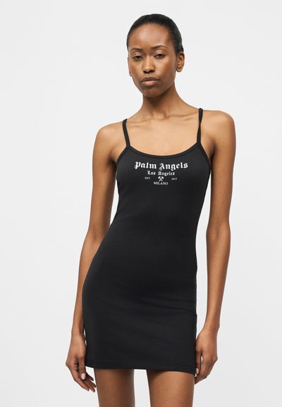 Woman wearing a black sleeveless mini dress with "Palm Angels Los Angeles Milano" text on the front, standing against a plain white background.