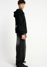 Czarna bluza z kapturem z kieszenią z przodu, z białym napisem na plecach. W zestawie z luźnymi, ciemnoszarymi dżinsami i czarnymi butami.