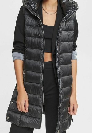 Persoon die een lange zwarte gewatteerde mouwloze jas draagt over een zwarte cropped top en een jas met opgerolde mouwen.