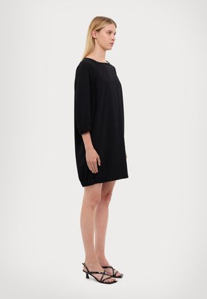 Schwarzes, oversized Kleid mit drei Viertelärmeln, das einen runden Halsausschnitt und geraffte Manschetten hat, getragen mit schwarzen, schlichten High Heels.