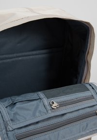 Jasno-brązowe wnętrze plecaka z ciemnoniebieską przegrodą, z zamkami błyskawicznymi, siateczkową kieszenią i gładkim materiałem. Uporządkowany i przestronny design.