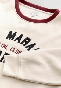 Sudadera color crema con cuello de canalé burdeos. Presenta gráficos de texto en negro y burdeos. Textura suave con mangas ajustadas.