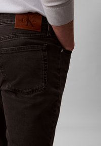 Dark denim jeans feature a close-fitting design, five-pocket style, and a brown leather tag with "Calvin Klein" embossed.